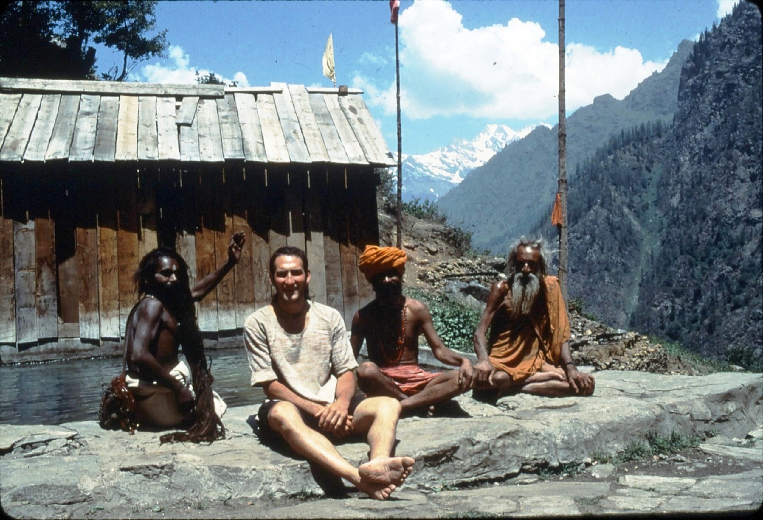 Full shot of a group of people, likely in a mountainous region.  Two men, dressed in traditional, orange-colored robes, are seated cross-legged on a rocky outcrop. One man has a prominent, long, flowing white beard.  A third man, with dark skin and long black hair, is seated beside the westernmost of the two men in orange, and is also barefoot.  A fourth man, light-skinned, wears a light-tan or beige-colored shirt and shorts. It is Rev Dr. Marlin Lavanhar. He is seated in front of the seated ones and is also barefoot.  Behind them is a small wooden structure with a simple, peaked roof of weathered, horizontal planks.  The background showcases a dramatic mountain range with snow-capped peaks, and lush green forested slopes.  A body of water is partially visible in the foreground, near the wooden structure.
