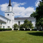 An image of All Souls Unitarian Church in Tulsa, Oklahoma, taken from Peoria Avenue. A classic, New England style church with a striking white exterior inhabits a vast, green campus.