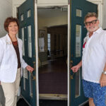 A woman and a man stand holding open a set of double doors leading into a hallway.