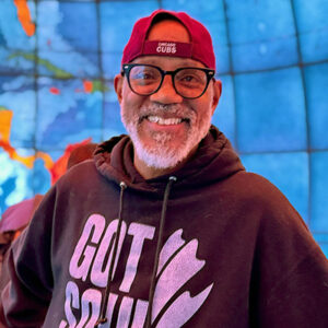 Corey Smith wearing glasses and an All Souls 'Got Soul?' hoodie with a Cubs hat, standing in front of a globe background.