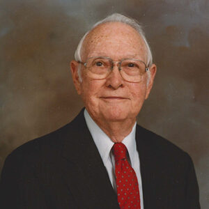 George Krumme, an older man in a dark suit, red tie, and glasses, smiling with a gentle expression.