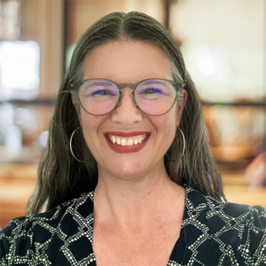 Hannah Middlebrook smiling, wearing glasses and a patterned top, with a blurred indoor background.