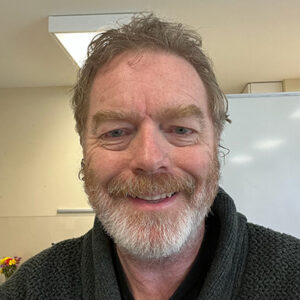 John Boston smiling, wearing a sweater, with a light indoor background.