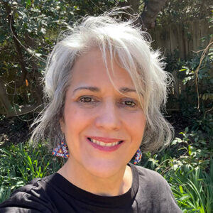 Lei Rumley smiling outdoors with white hair, wearing a black shirt, with greenery and a wooden fence in the background.