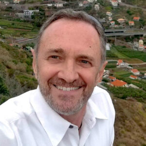 Leo De Andrade smiling outdoors with a scenic background of green hills and houses.