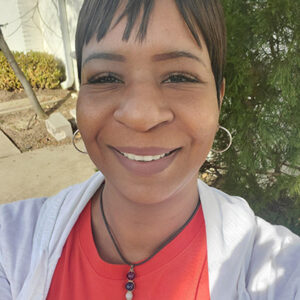 Nefertiti Sanders smiling outdoors in a red shirt and necklace, standing in front of a building and greenery.