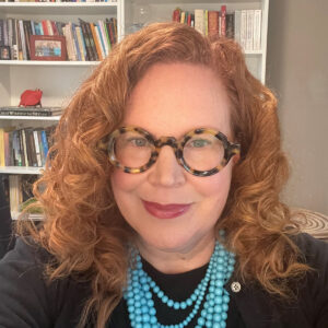 Rev. Dr. Nicole Kirk smiling, wearing round tortoise-shell glasses, blue necklace, and standing in front of bookshelves.