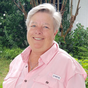 Shelley Mackin smiling outdoors in front of a white building and greenery.