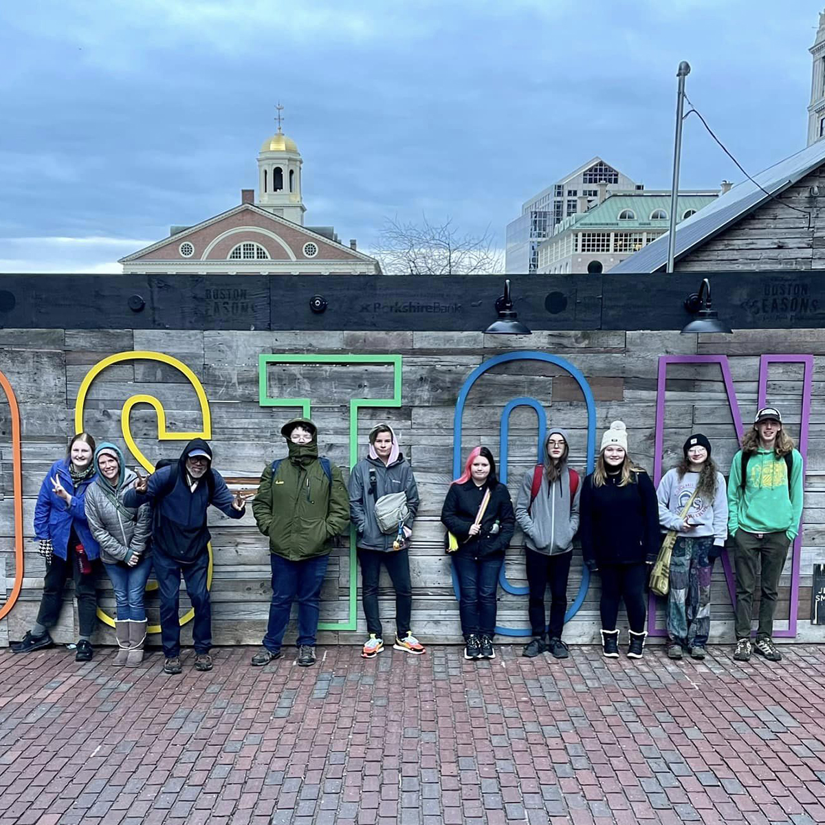 Coming of Age youth and leaders pose in front of colorful 'Boston' letters during their 2023 trip, celebrating their journey.