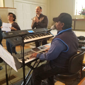 David Smith sits at a keyboard singing, with choir members and Rev. Marlin Lavanhar singing in the background.