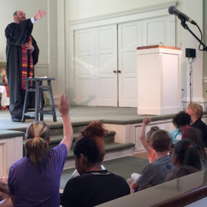 A minister in a robe addresses a group of children seated on the steps of the chancel during a service at All Souls Unitarian Church.