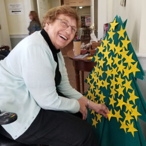 A smiling volunteer places stars with wishes on a green tree-shaped board during a community event at All Souls Unitarian Church.