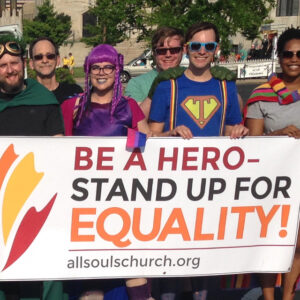 All Souls Unitarian Church members dressed as superheroes hold a banner that reads 'Be a Hero—Stand Up for Equality!' at a Pride event.