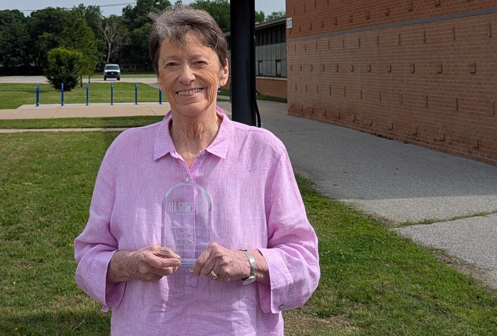 Margo Mitchell stands outdoors on a sunny day, smiling warmly at the camera. She is wearing a light pink button-up shirt and holding a clear glass award in front of her with both hands. The award is engraved with the All Souls logo and text that is partially visible. Behind her is a brick building with a sidewalk and grassy area, and in the distance, a white van is parked near some trees. The setting appears to be a school or community space.