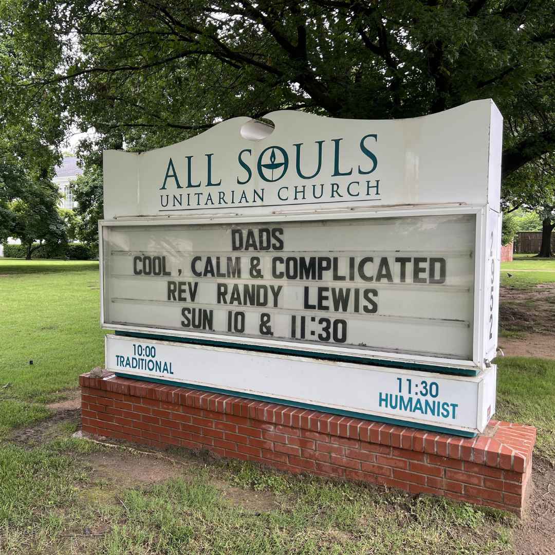 An outdoor sign for **All Souls Unitarian Church** stands on a small red brick base, surrounded by lush green grass and trees. The message on the marquee reads: **“DADS** **COOL, CALM & COMPLICATED** **REV RANDY LEWIS** **SUN 10 & 11:30”** Beneath the main message, smaller blue text indicates the types of services: “10:00 TRADITIONAL” on the left and “11:30 HUMANIST” on the right. The sign is situated under a canopy of trees, giving a peaceful and welcoming feel.